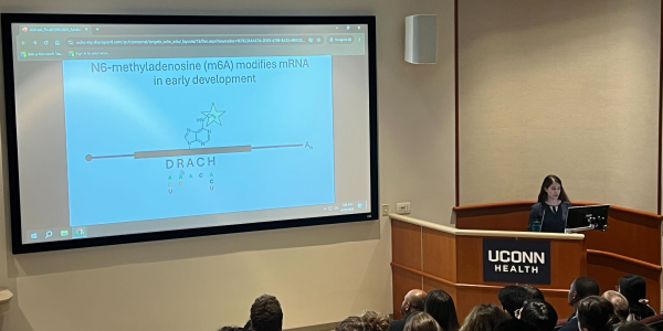 photo from back of auditorium of a student in right corner of image standing behind a podium with a sign reading "UConn Health" with her presentation on a large screen to her right