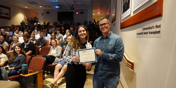 Osborn Award winner Pedro Miura standing on the botttom steps in an auditorium to the right of a student presenting him with an award certificate with audience members sitting in the background