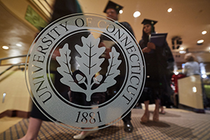 UConn seal on a glass window with graduates walking behind it