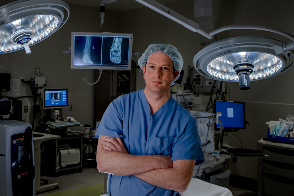 Adam Lindsay, MD, standing in the operating room with an X-ray of a foot on a monitor in the background