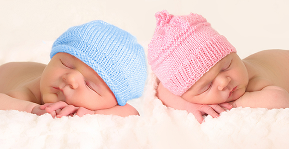 Two babies laying side by side, one wearing a blue hat an one with pink
