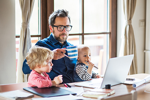 A father sitting at the table working, with daughter sitting on his lap and son watching